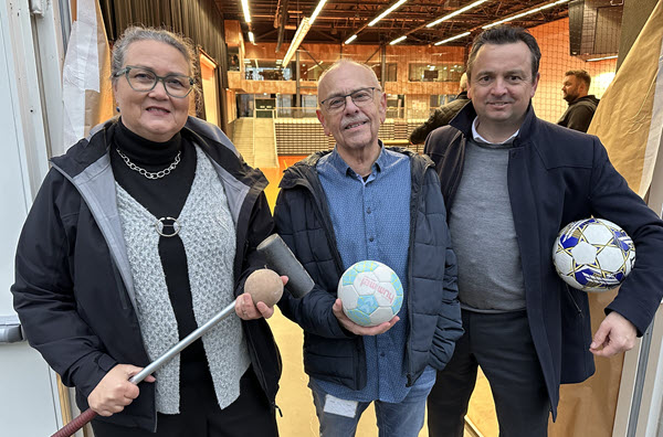 Tre personer står foran byggepapir i døråbningen til en hal og har henholdsvis en fodbold, en håndbold og et krolfsæt i hænderne.